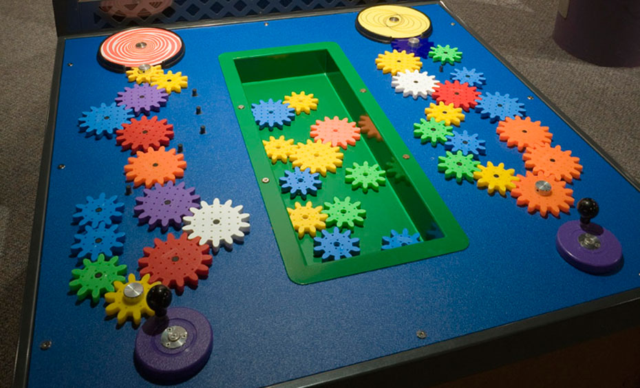 Interlock rows of different sized gears and make an illusoin disc spin. On blue and black table top, sits multiple different coloured and sized gears, interlinked and sitting in a green tub.
