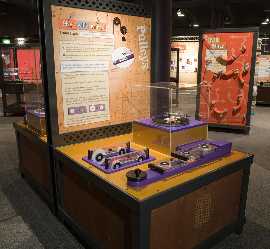The size and combination of pulleys determines how fast they (and things that are attached) will spin. A timber, black steel and yellow exhibit table with a matching vertical information panel at the back. On the table top sits a record player in a perspex box surrounded by purple and black gears contained in rectangular perspex boxes.