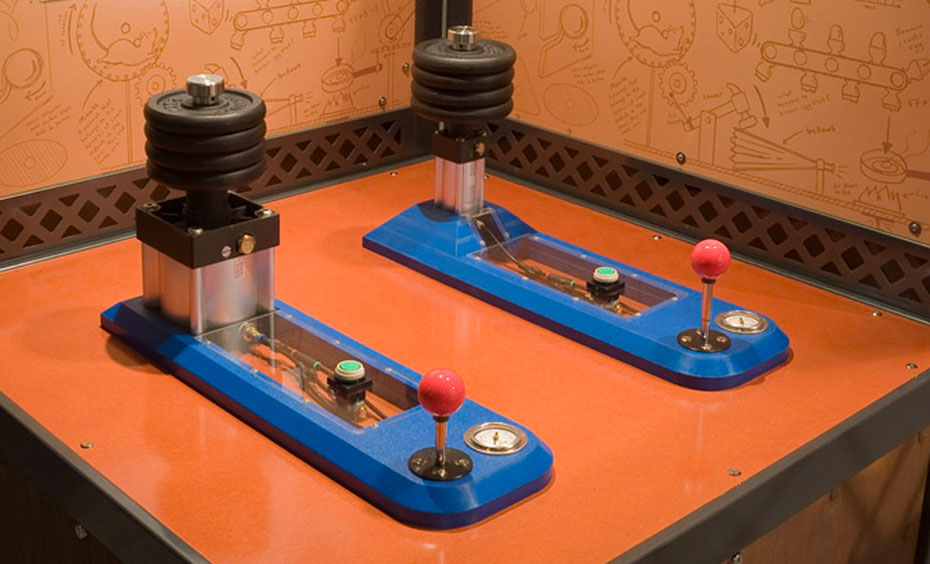One narrow and one wide piston are pumped up to lift masses to a certain height. A timber, black and orange exhibit table with two information panels at the rear in the same colour scheme. On the table top sits two piston devices with blue bases and a series of weights at one end. Thre is a red joystick and pressure guage at the other end.
