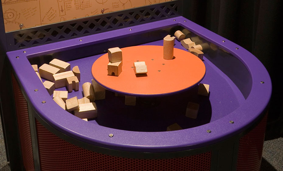 Children under 6 years build with wooden blocks on a fixed or unstable tabletop and experiment with centre of mass. A large purple tray containing wooden building blocks, has a raised orange round platform in the centre of it.