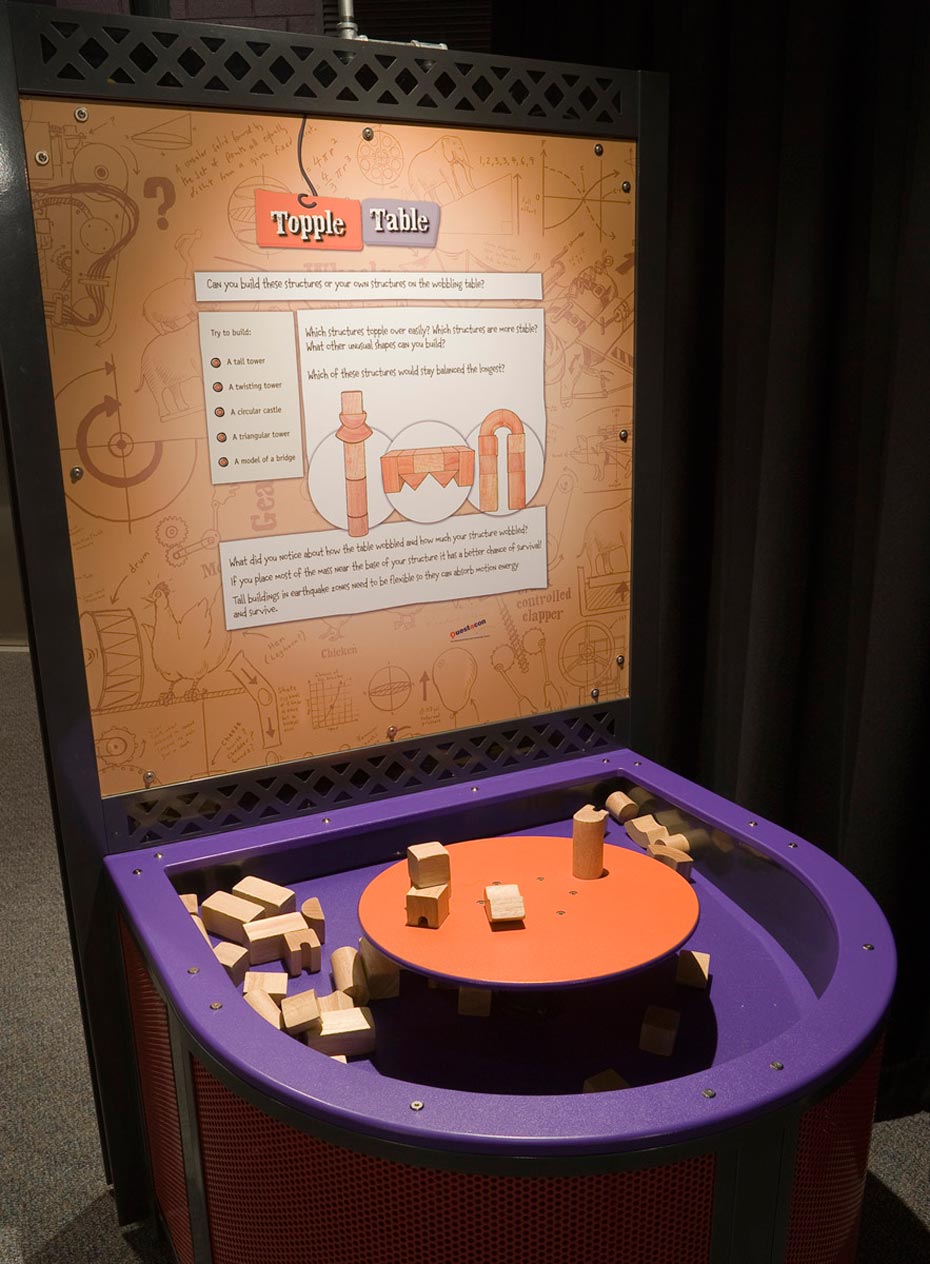 Centre of mass creates stability within a structure. A large purple tray containing wooden building blocks, has a raised orange round platform in the centre of it. At the back of the purple tray is a brown and white information panel.