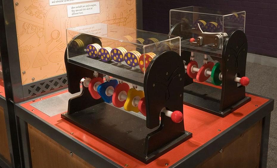 A toy caterpillar and toy dog have rods attached to parts of their body, so different camshafts make them ‘jump’ or ‘wriggle’ in different ways. A timber, black and red exhibit table with matching information panel at the back in brown and white has two toy engines on the table top. The engines show the cam shaft and pistons, and are brightly coloured.