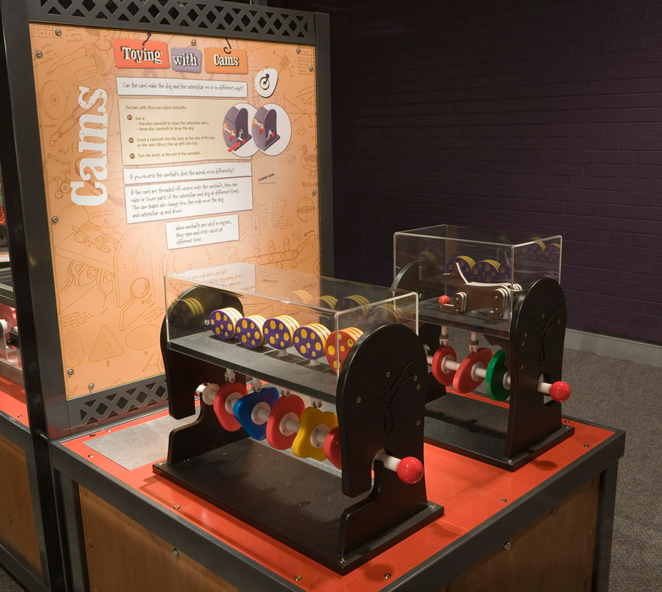 The shape of a cam disc influences the pattern and timing of how other things move. A timber, black and red exhibit table with matching information panel at the back in brown and white has two toy engines on the table top. The engines show the cam shaft and pistons, and are brightly coloured.