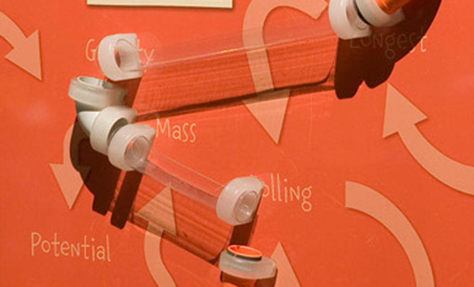 Line up tubes on the walls and roll a ball through the tubes. An orange vertical surface, with clear plastic piping attached. the words 'potential', 'mass', and 'longest' are displayed along with arrows pointing in every direction.