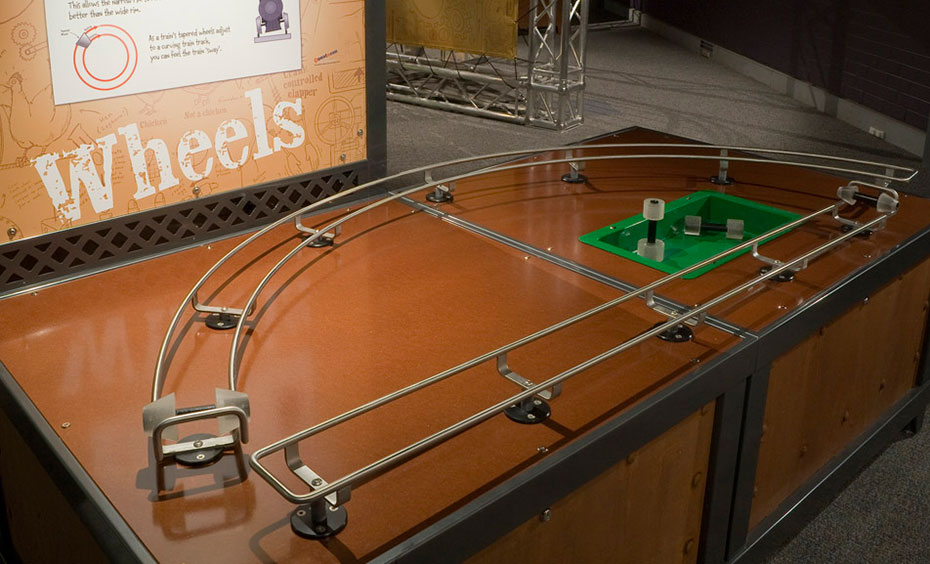 Roll sets of tapered wheels along a track to demonstrate how train wheels roll along train tracks. A brown and black exhibit table with matching information panel at the back. On the table is a D shaped rail with wheels sitting on the track.