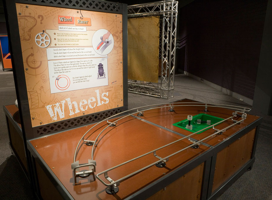The shape of wheels determines how well they follow different shaped tracks. A brown and black exhibit table with matching information panel at the back. On the table is a D shaped rail with wheels sitting on the track.