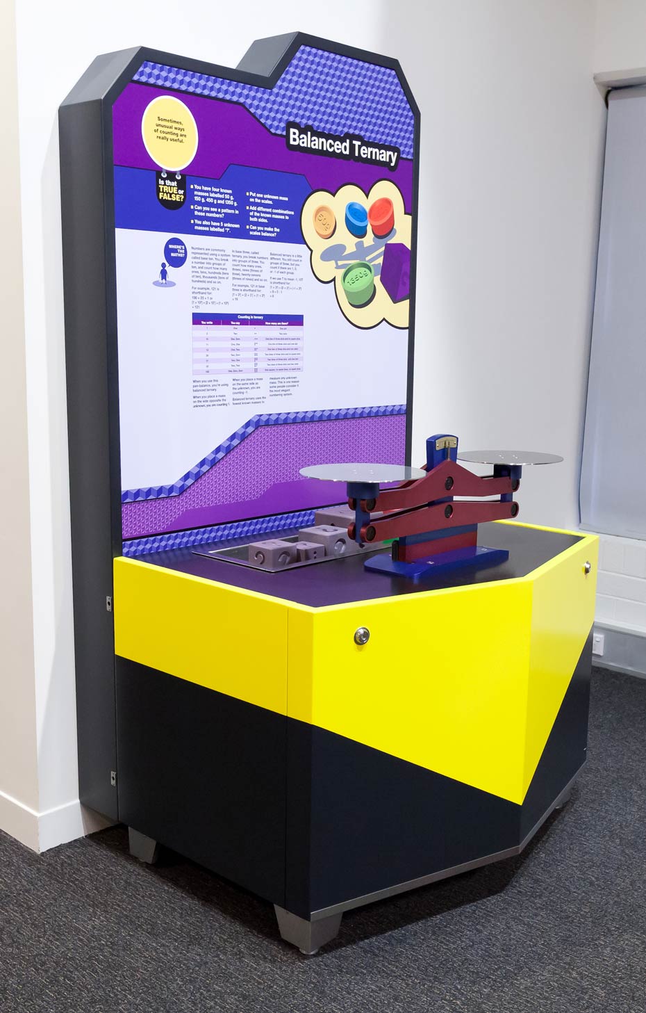 The ternary counting system is based on 'threes', while our traditional decimal counting system is based on 'tens'. A black, yellow, white and purple exhibit sitting in a white room. On the exhibit table sits a blue and red scale.