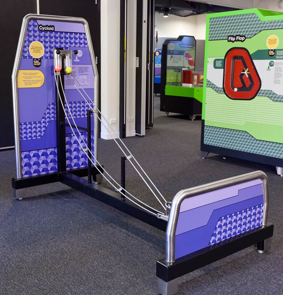 Race two balls down two separate tracks: one straight track (which appears to be shorter) and one curved or cycloid track (which appears to be longer). A tall and short purple vertical panels joined together by black steel bars and two metal ball tracks.