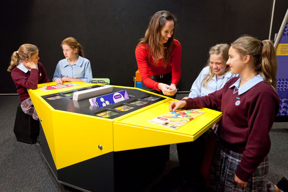 Sixty different maths puzzles have been printed onto placemat-sized panels. A wowan and four school girls are sitting next to a yellow and black table that has an assortment of maths puzzles on top.