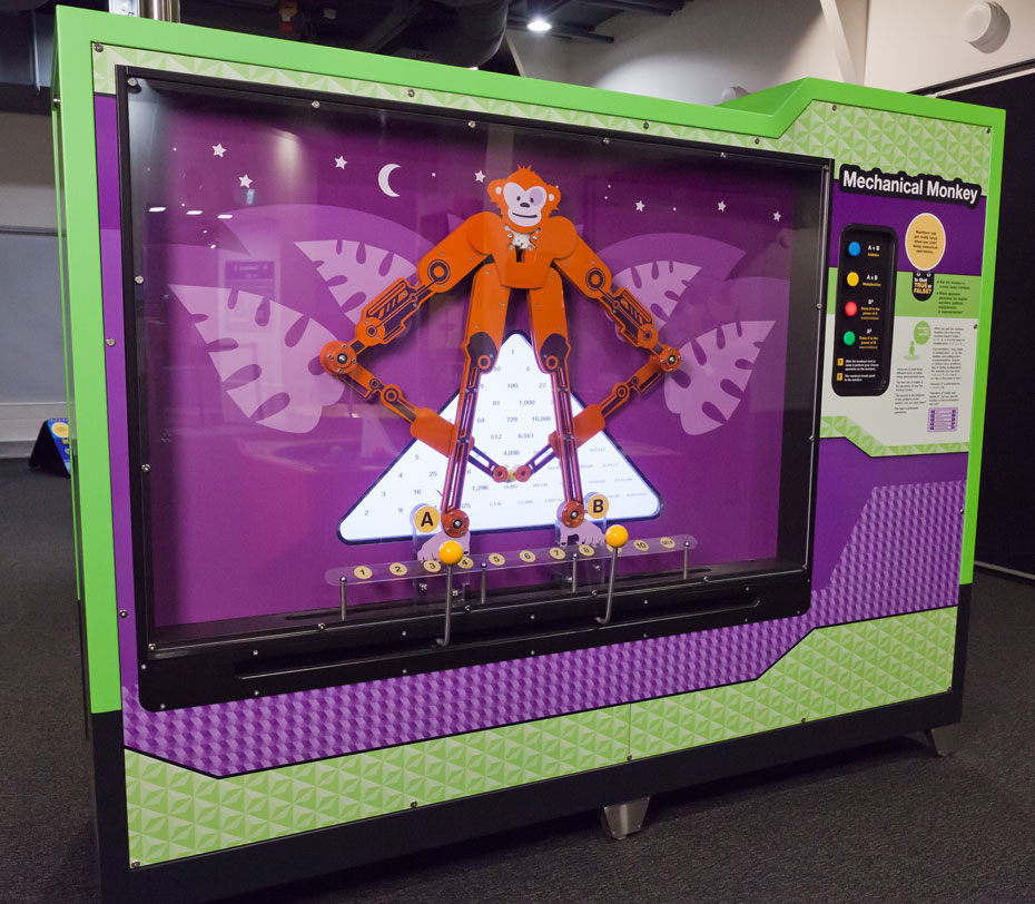 Certain numerical operations create much larger numbers than other numerical operations. A large rectangular green and purple exhibit with an orange monkey on the left behind glass and coloured buttons on the right information panel.