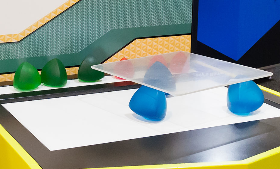 Roll a square plate on top of different-shaped solids, and feel which shapes allow the plate to roll smoothly. A yellow and black exhibit table and information panel. On the table top sits blue and green solid shapes and a square glass plate.
