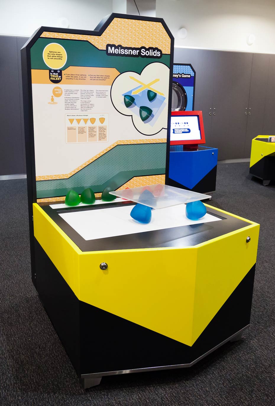 Can you find other shapes that will allow the plate to roll just as smoothly? A yellow and black exhibit table and information panel. On the table top sits blue and green solid shapes and a square glass plate.
