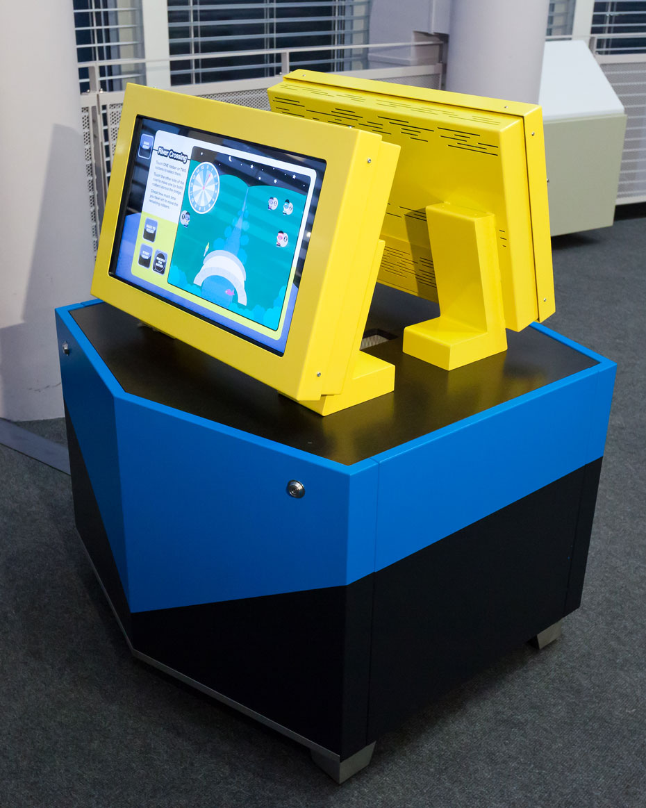 Use the touch screen and certain puzzle rules to move four robbers across a bridge to the opposite river bank within a '15 minute' time limit. Two yellow monitors back to back sit on a black and blue exhibit table.