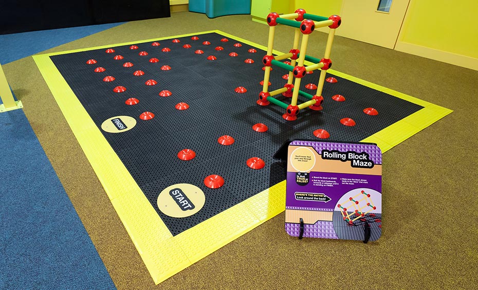 Roll a giant rectangular prism puzzle piece along the floor maze pathway from 'start' to 'finish'. A large black rubber mat with a yellow border and red points throughout the black area has a yellow and red plastic rectangle frame sitting on four of the red points.