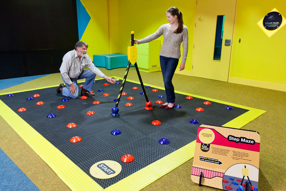 Can you find the path through this maze? A man and a young woman are standing on a black mat with a yellow border and red and blue markers throughout. The woman is holding a large black and yellow divider.