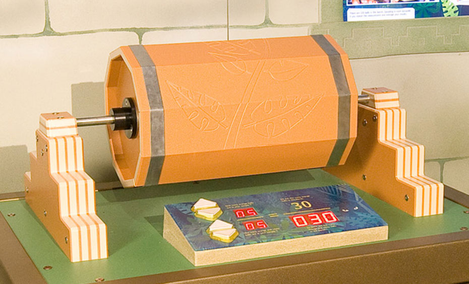 Capture and count balls in a barrel to model estimation of animal populations. A fake stone wall in the background with a blue information panel displayed. In the foreground is a brown table with an orange cylinder on top laying on it's side with a steel spindle through it's centre. There are controls in front of this on the top of the table.