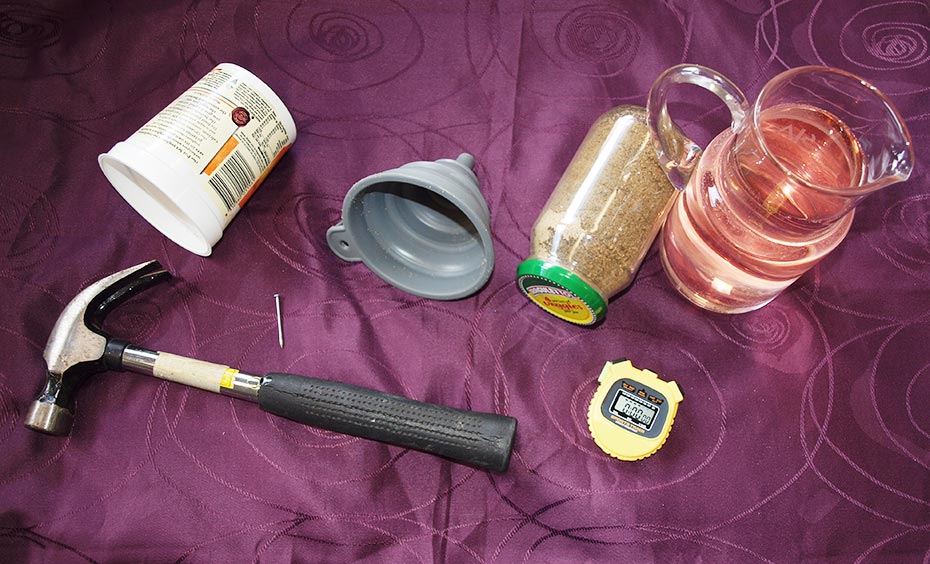 A maroon coloured table cloth with a hammer, plastic cup, nail, grey plastic funnel, glass jar of sand with a green and yellow lid, yellow stop watch and glass pitcher of apricot coloured water siting on it.
