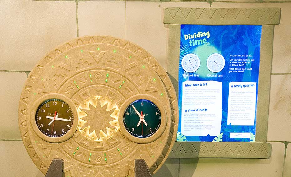 Compare a standard (12 hour) clock with a decimal clock (10 hours) and try to assess the time of day using each clock. A fake stone wall with a blue information panel in the background. In the foreground is a brown and green table, with a large vertical ciruclar disc with different shapes etched into it. On the face of this disc are two clocks.