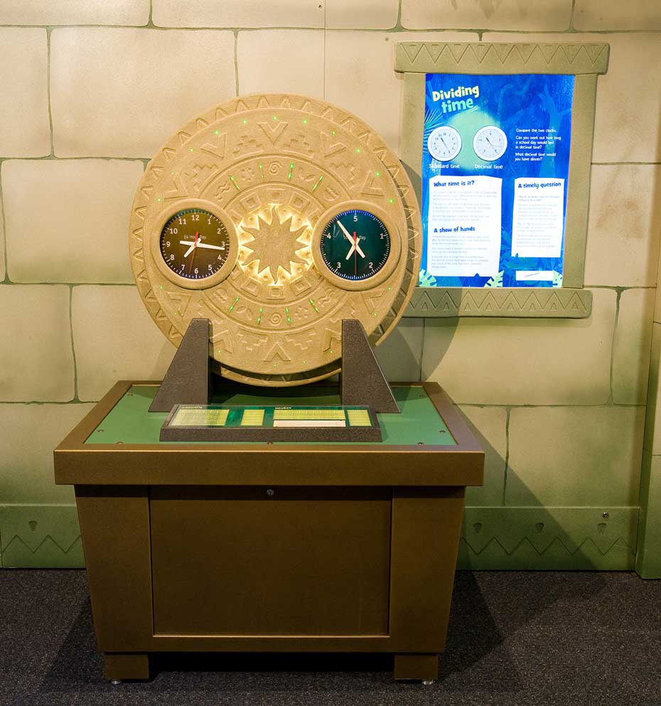 Standard time clocks are based on 12 hours, but time can also be measured using a decimal clock based on 10 hours. A fake stone wall with a blue information panel in the background. In the foreground is a brown and green table, with a large vertical ciruclar disc with different shapes etched into it. On the face of this disc are two clocks.