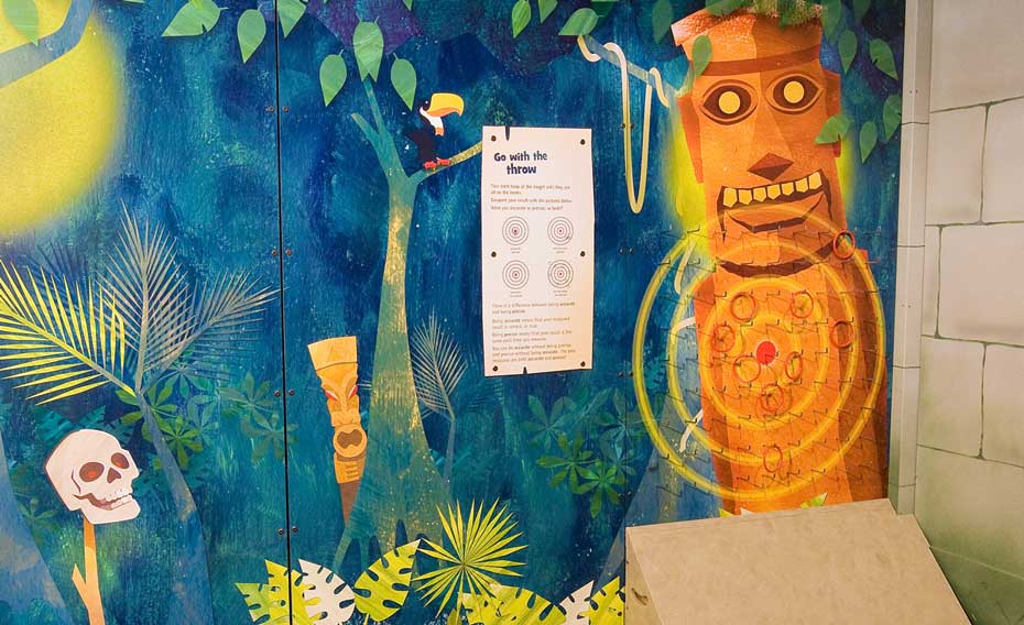 Throw quoits at a target and work out whether you were accurate, precise, or both! A wall mural painted as a jungle scene in blue, green and orange, with pictures of a skull, a mask, a bird, a information panel and a totem pole that has a target on it.