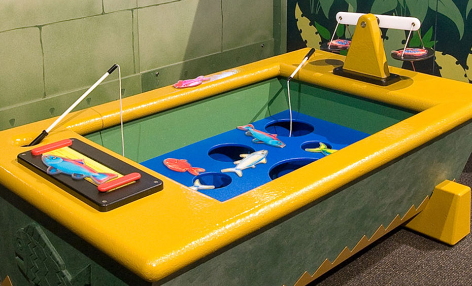 Catch two toy fish and compare the fish on a ruler and a set of weighing scales. A yellow and blue tub with cut out fish and small fishing poles sits in front of a fake stone wall and jungle scene.