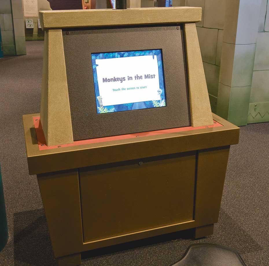 Touch monkeys on the screen that are hidden within a misty jungle and check your score representing how well you can detect grey shades overlaid onto darker or lighter backgrounds. A brown exhibit table with a vertical dispaly area that has a monitor screen and the words 'Monkeys in the Mist' on the screen.