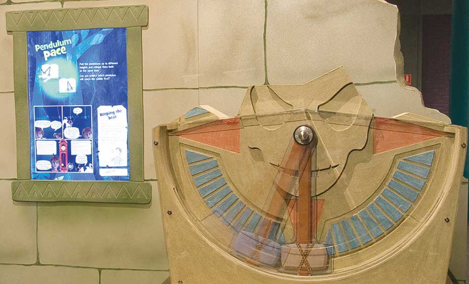 Swing two pendulums from different heights and estimate which pendulum will reach the centre first. A fake stone wall with a blue and white information panel on it, sits behind a brown exhibit table with a vertical pendulum siting on top.