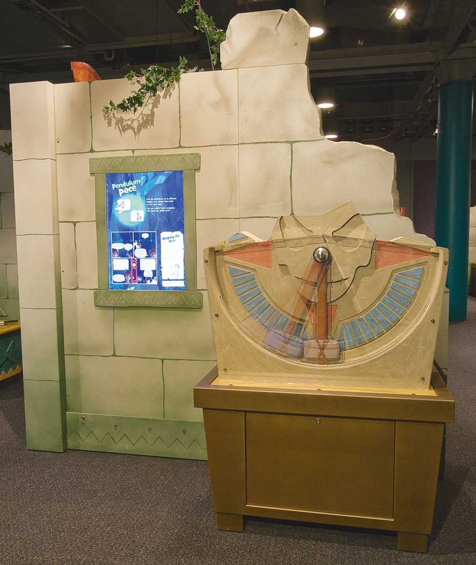 Pendulums are used to keep time in certain clocks, based on their properties of length and patterns of swing. A fake stone wall with a blue and white information panel on it, sits behind a brown exhibit table with a vertical pendulum siting on top.