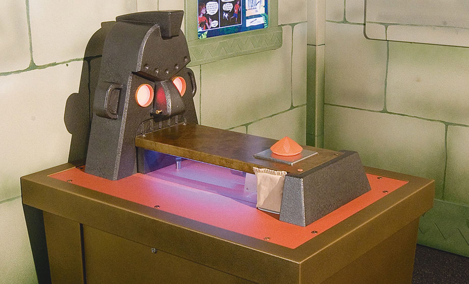 Swap a 'diamond' sitting on a pressure sensor with a bag of sand, so you don't wake the statue. A brown exhibit table with a steel mask with glowing eyes sitting on top, and a rectangular platform coming out of the statue's mouth. In the background is a fake stone wall with a blue, white and green information panel.