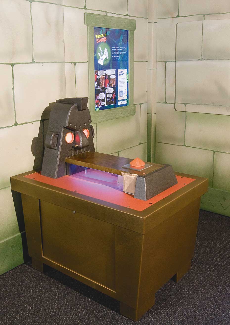 Sensors detect force exerted by a weighted object. A brown exhibit table with a steel mask with glowing eyes sitting on top, and a rectangular platform coming out of the statue's mouth. In the background is a fake stone wall with a blue, white and green information panel.