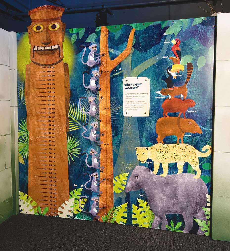 Young children can measure their height against a ruler, or a stack of monkeys and other animals. A colourful wall mural with a large totem pole on the left that has measurement markings in centremetres from top to bottom. The mural also has monkeys in a tree and various animals stacked upon one another from large to small.