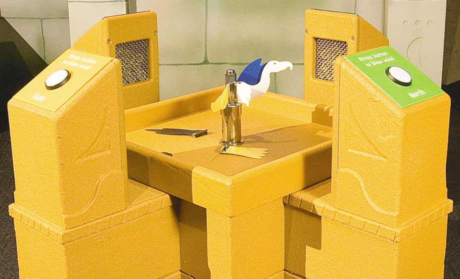 Construct a wind vane by attaching parts to a bird that is surrounded by air blowers. An unusually shaped yellow exhibit table, that has a button to press at each of the four corners and has a model bird in the centre.