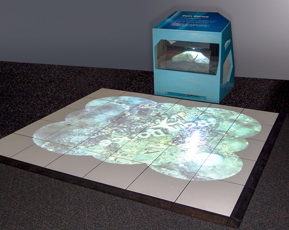 When raindrops congregate in a cloud to a certain density, rain will fall. White tiles on the floor have circular light projection onto them, from a blue box at the back of the tiles.