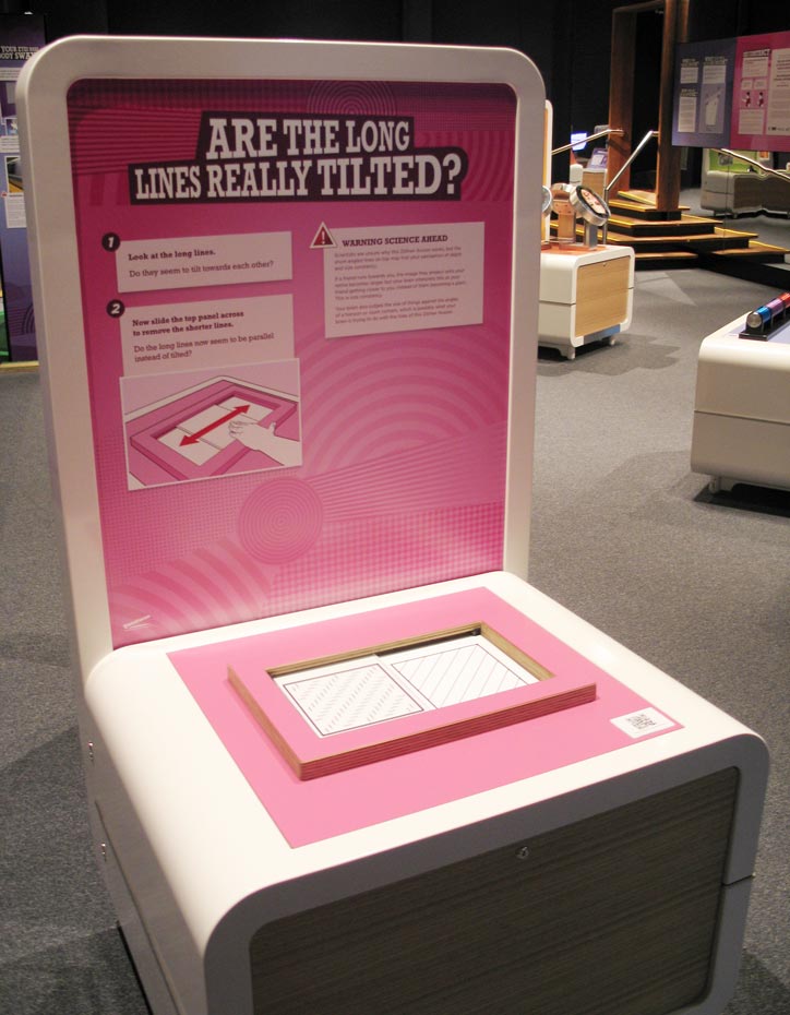 Slide a panel of short angled lines across a panel of long lines and notice whether the long lines change in appearance. A cream and pink exhibit table and display board with the title 'Are the long lines really titled?'. Two squares with diagonal lines appear on the table top.