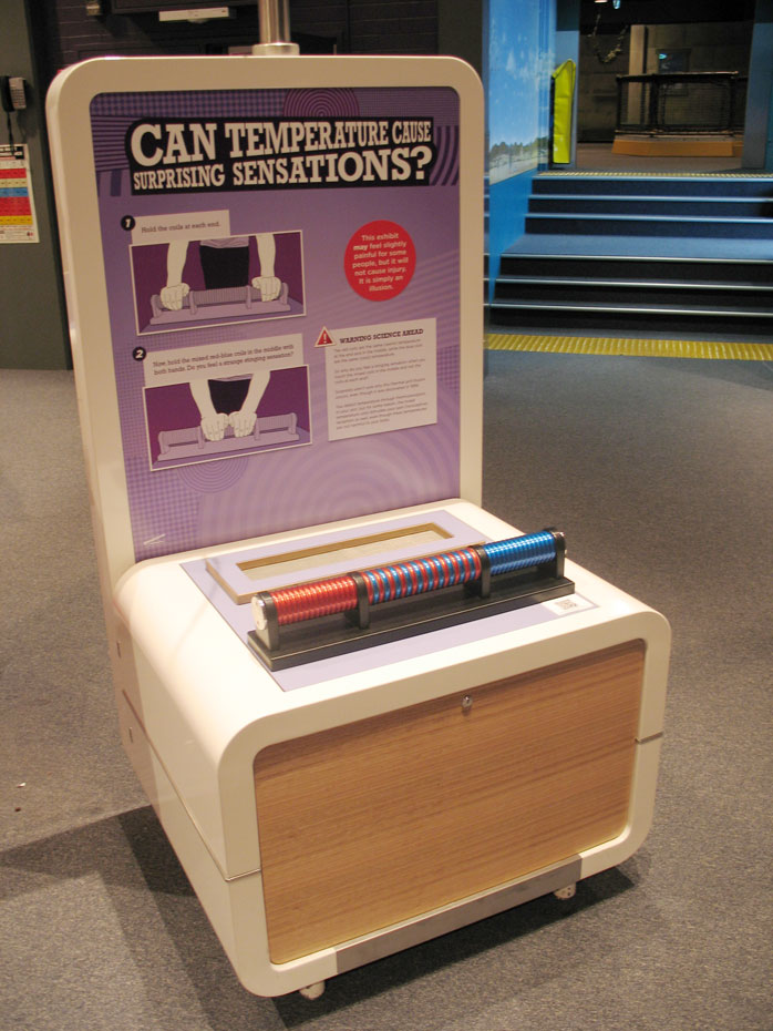 The thermal grill tactile illusion uses mild temperatures to create an illusion of discomfort. An exhibit table and headboard with the text 'Can Temperature Cause Surprising Sensations?'. There are a row of discs on the table top.