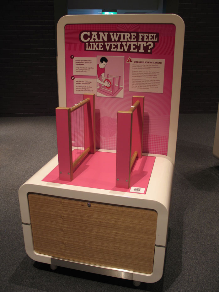 The tactile Velvet Hand Illusion (VHI) demonstrates how the brain can 'fill in' different types of information. An exhibit table and display board, with vertically strung wires on timber frames. The title 'can wire feel like velvet?' appears on the backboard.
