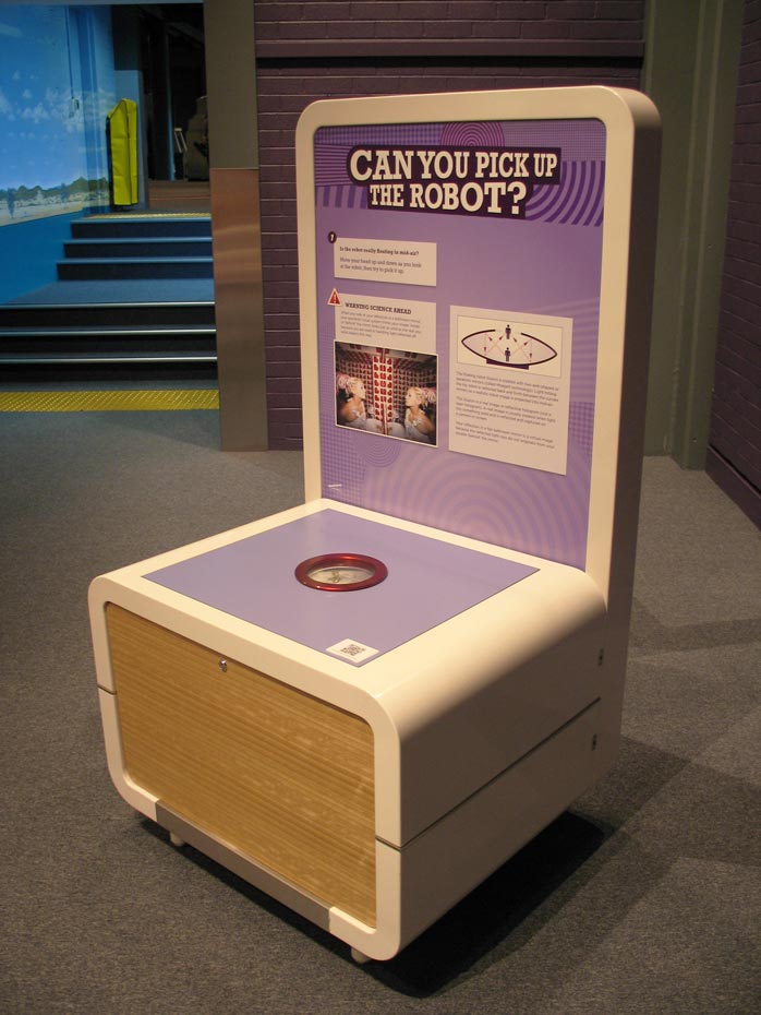 Reflect or mirage holograms use specially-curved mirrors to make objects appear to float in mid-air. A cream and purple coloured exhibit table with headboard that has 'Can you pick up the robot?' written upon it.