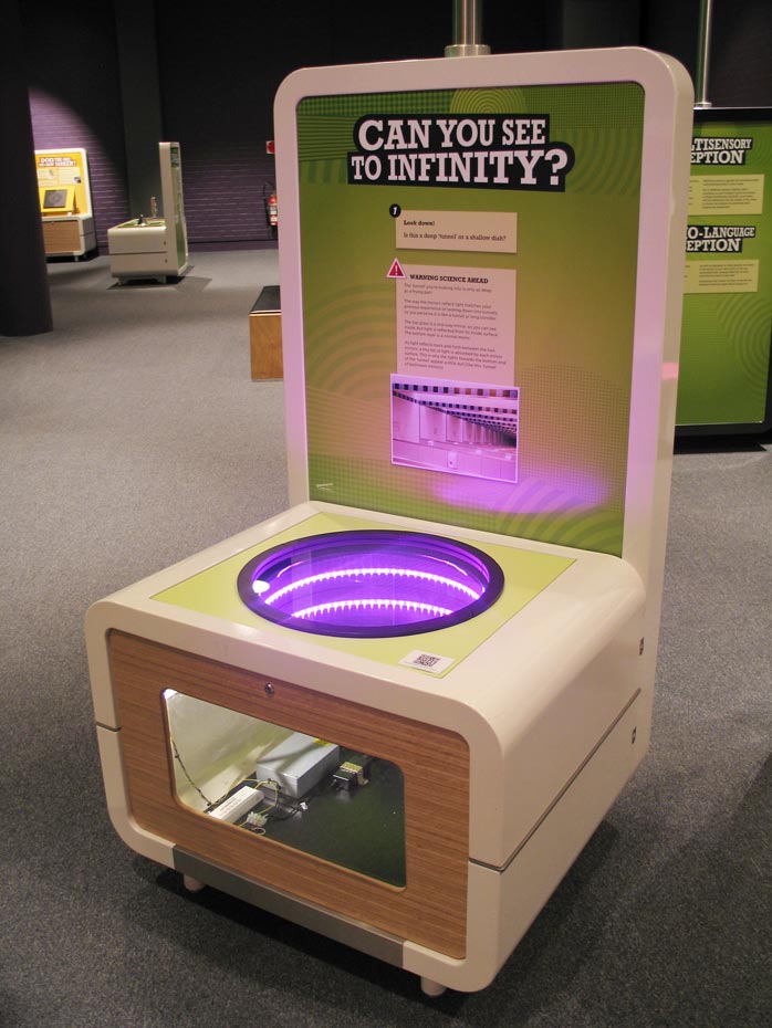 Look into a round hole and see a ‘tunnel’ burrowing deep into the ground, even though the hole is about as deep as a frypan. A cream and green coloured exhibit table and backboard with the title 'Can you see to infinity?' on it. On the top of the table is a blue glass hole with lights.