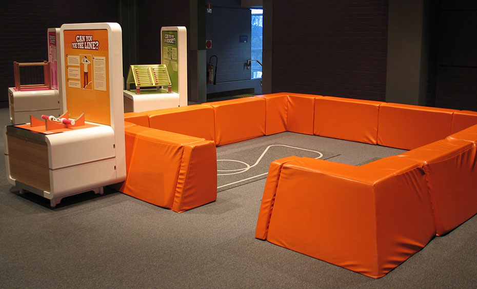 Hold a special pair of prism glasses up to your eyes and try to walk around looping patterns on the ground. When you look through the prism glasses, your view of the world becomes skewed, as does your sense of balance. A cream and orange exhibit table stands in front of orange knee high padded fences that re sitting on brown-grey carpet.