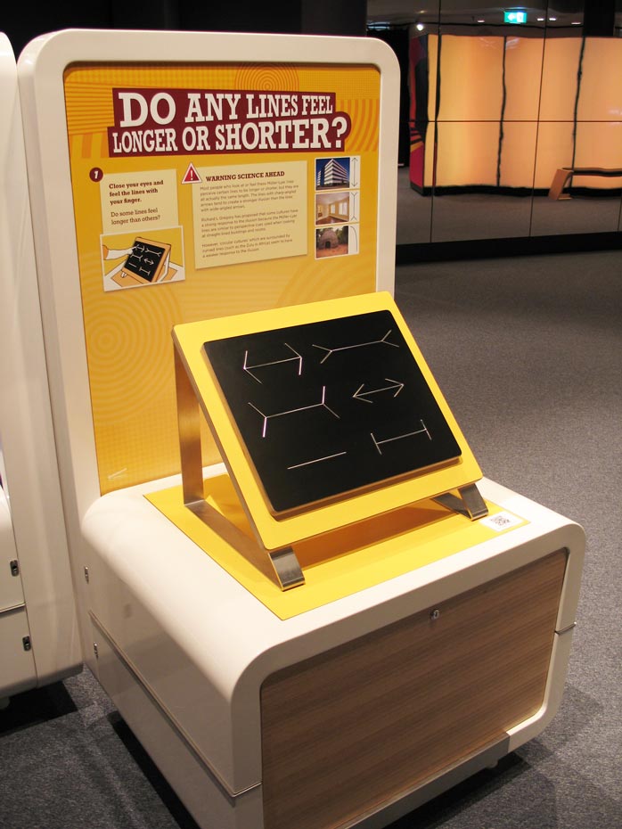 This is a tactile version of the Müller-Lyer line illusion. Close your eyes and feel six different raised lines on the board. Even though all of the lines are the same length, some will feel longer or shorter than others (similar to when you simply look at the lines). A cream and yellow exhibit table and headboard with the title 'do any lines feel longer or shorter?'. on the table top is a yellow and black board with metal lines that have arrows, no arrows or perpendicular lines on each line length.
