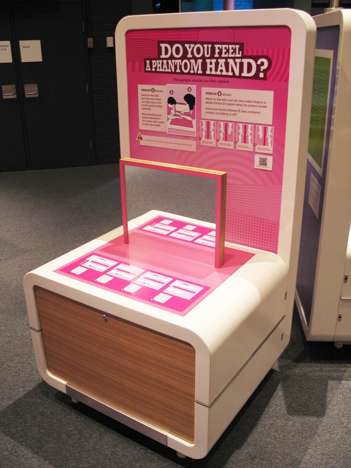 This exhibit requires two people. One person will place their hands on either side of a mirror and keep watching their hand's reflection in the mirror. The second person will brush or stroke the other person’s palms in a particular pattern, so the other person feels their hand being touched, but not see anything touch their hand! A cream and pink exhibit table and back board that has a vertical mirror on the table and the words 'do you feel a phantom hand?' on the backboard.