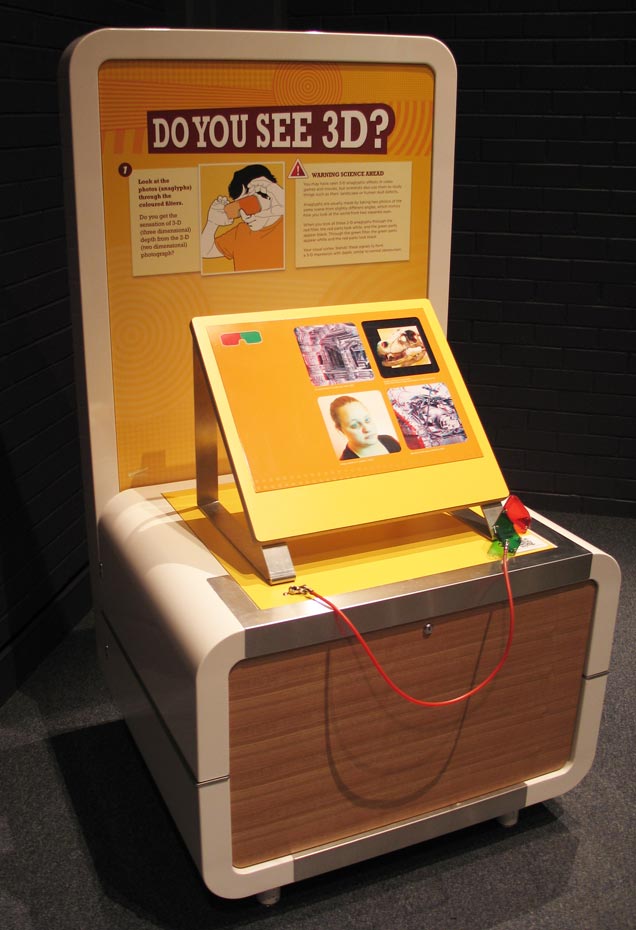Look at special photos through coloured filters, so the photos appear to be three dimensional (3D). A cream, orange and yellow exhibit table and backboard, with the title 'Do you see 3D?
