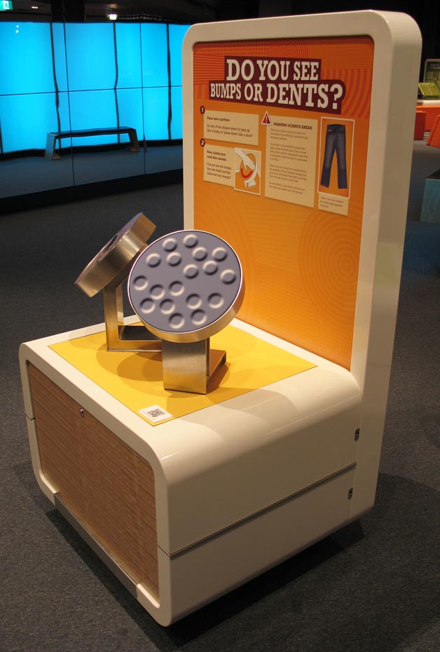 What happens if we leave the disc alone, but we turn out heads upside down and look at the pictures? A cream, orange and yellow exhibit table and backboard, wit the words 'Do you see bumps or dents?'. On the table sits two round displays that have grey bumps and dents on the surface.
