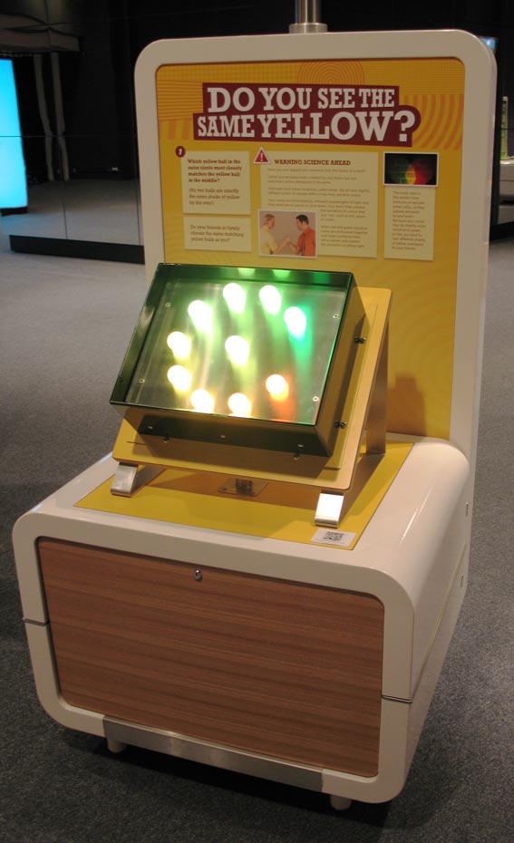 Look at a circle of ping pong balls illuminated by LEDs (light emitting diodes) so they appear ‘yellowish’. The middle ping pong ball contains pure yellow LEDs, while ping pong balls in the surrounding circle have different proportions of red and green LEDs to give yellowish coloured light. A cream and yellow exhibit table and headboard with the title 'Do you see the same yellow?'. A black box with 10 lights in a circle sit on the table.