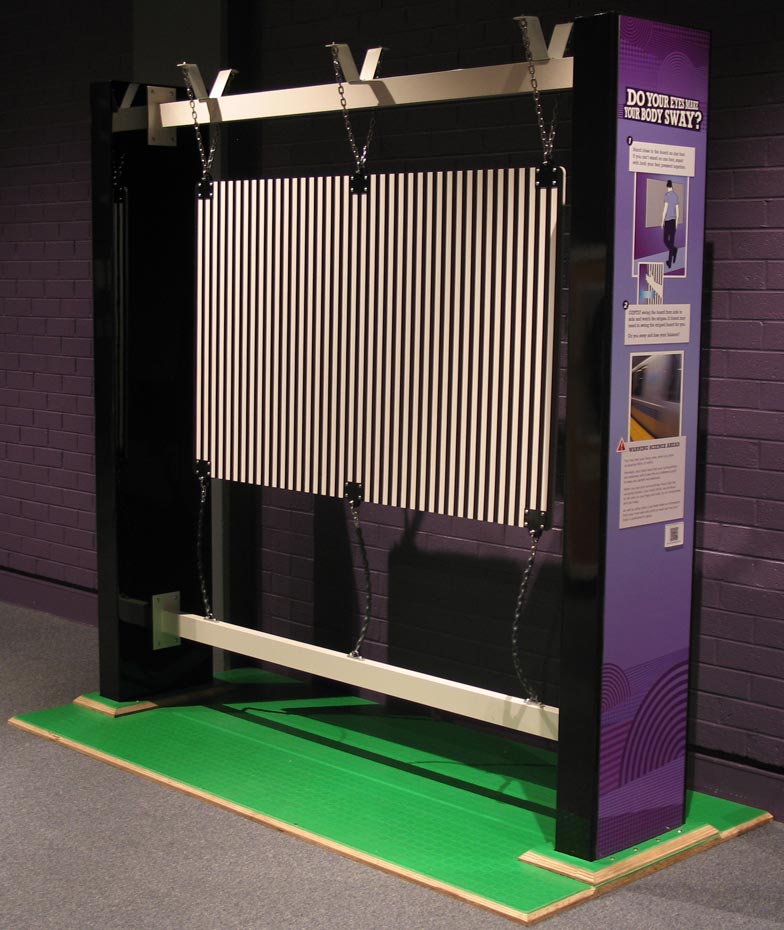Stand on one leg, up close to a swaying board printed with black and white stripes. Can you keep your balance? A large pruple and cream coloured vertical exhibit, that has a series of vertical timber pieces hung between two large panels.