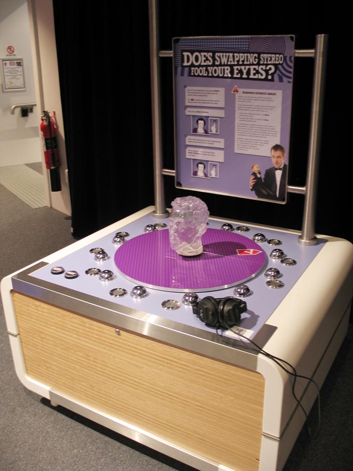 Wear a pair of headphones while you watch and listen to ringing bells around a dummy's head. The headphones let you listen to the ringing bells from the perspective of the dummy's head. A cream and purple exhibit table with back board that has the text 'Does swapping stereo fool your eyes?' displayed. A clear plastic head sits on a purple flat disc along with black headphones on the table top.
