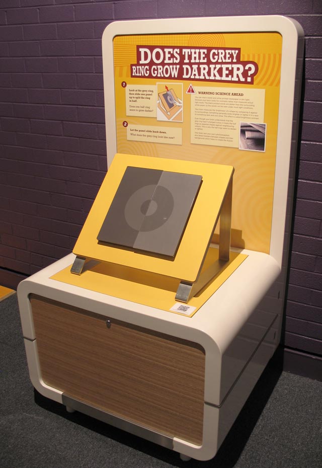 A grey ring (called a Koffka ring), is printed across two panels. When you slide one panel upwards, the grey ring splits in half and it seems to change shade. The ring’s shade doesn’t change, only your perception of its colour based on its surroundings. A cream and yellow exhibit table and headboard with the title 'Does the grey ring grow darker?'. A grey square and circle sit on the table top at 45 degrees.