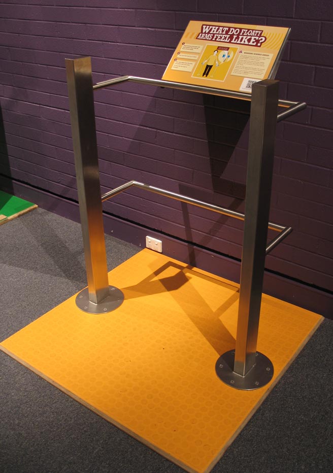 This old trick (discovered in 1915) shows how your brain and muscles constantly respond and adjust to incoming signals. A yellow sign with the title 'what do floaty arms feel like?' supported by a stainless steel frame that is bolted to a yellow floor mat.
