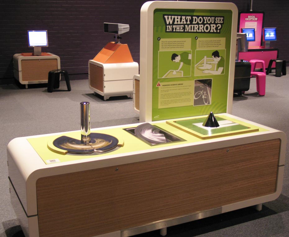 Place some puzzle pieces around a column-shaped or cone-shaped mirror. The puzzle pieces seem to look smeared or warped, but when you put the pieces in place and look at the mirror, you’ll see something more recognisable. A cream and green coloured exhibit table and vertical information panel with the wording 'What do you see in the mirror?'. On the table are a chrome vertical short pole, a half circle and a black small inverted cone.