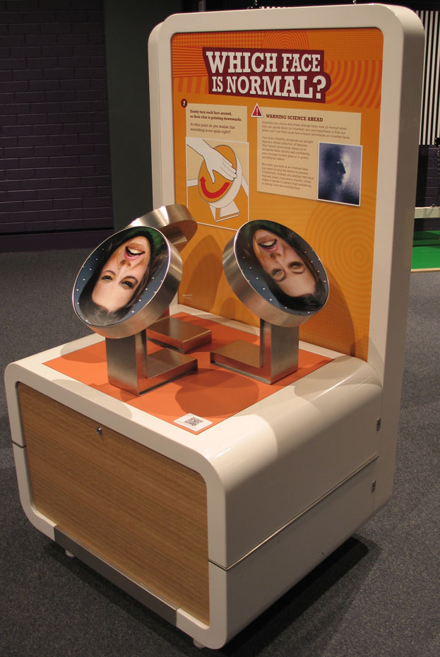 Digitally altered photos of faces are printed upside down on discs. Look at each inverted face, then turn the discs around to see the face in their normal upright position. A woman's face printed onto a round steel disc, sits on an exhibition table
